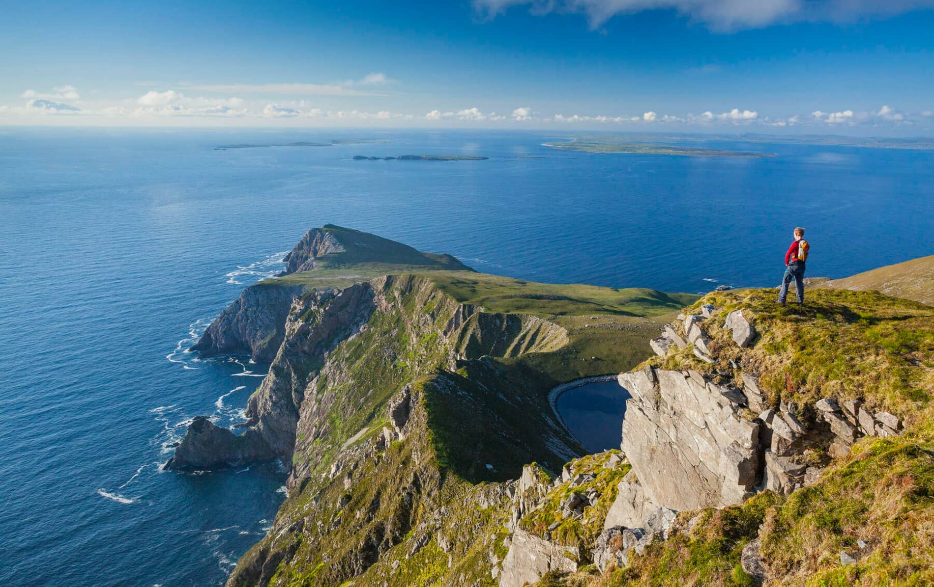 Person in red jacket standing on a cliff edge on Achill Island, overlooking the Atlantic Ocean on a sunny day.