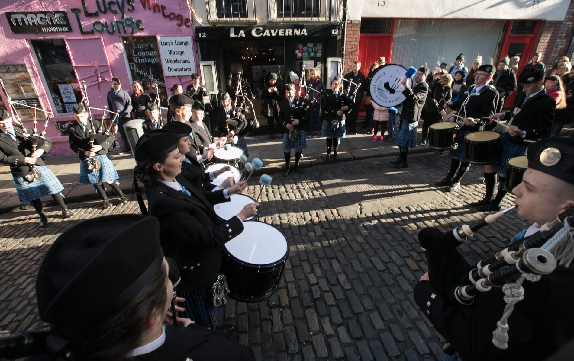 Pipe band musicians perform on a cobbled street in Temple Bar, Dublin, watched by a crowd in winter clothing.