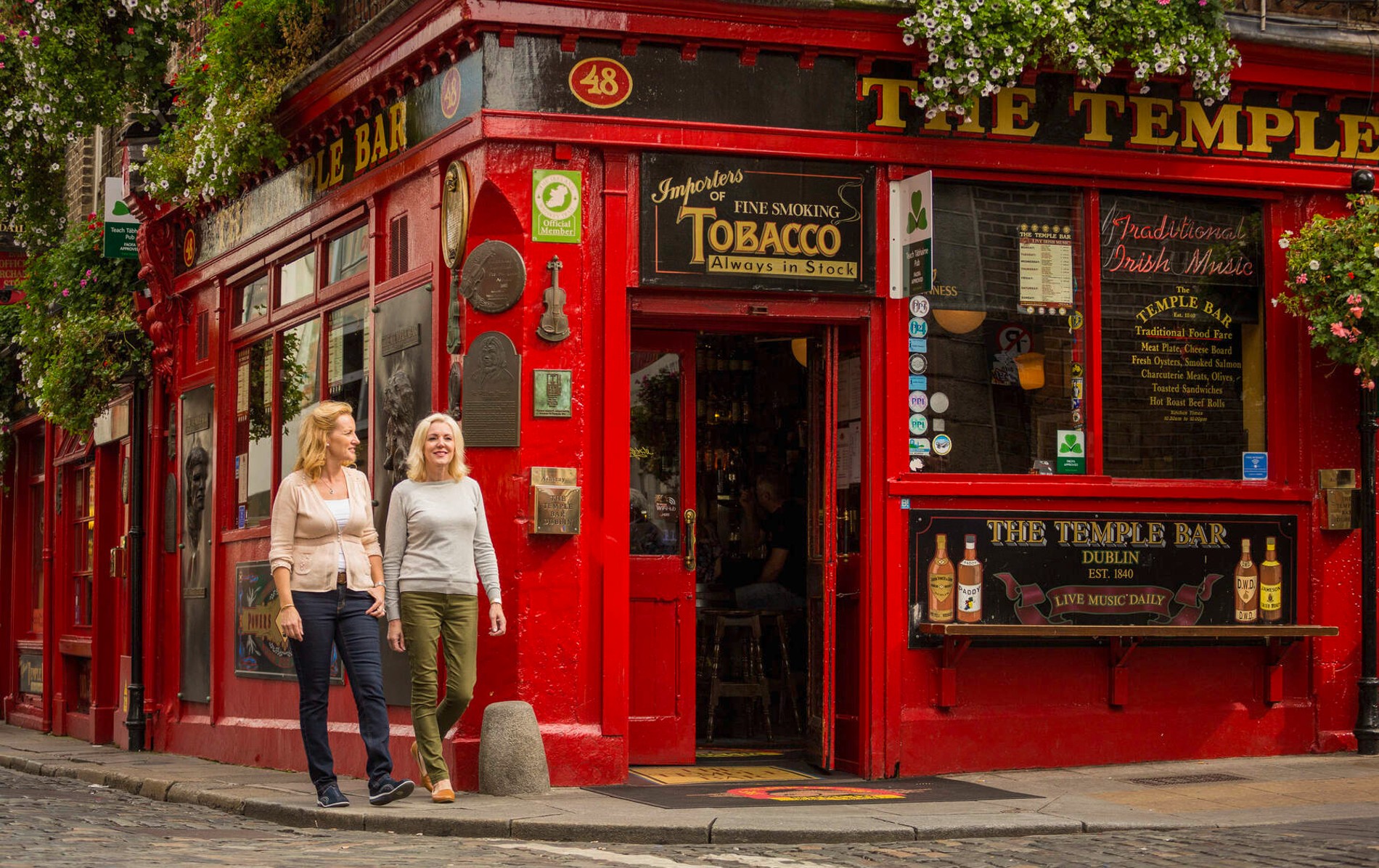 Two women walk past the iconic red façade of The Temple Bar pub in Dublin, known for traditional Irish music.