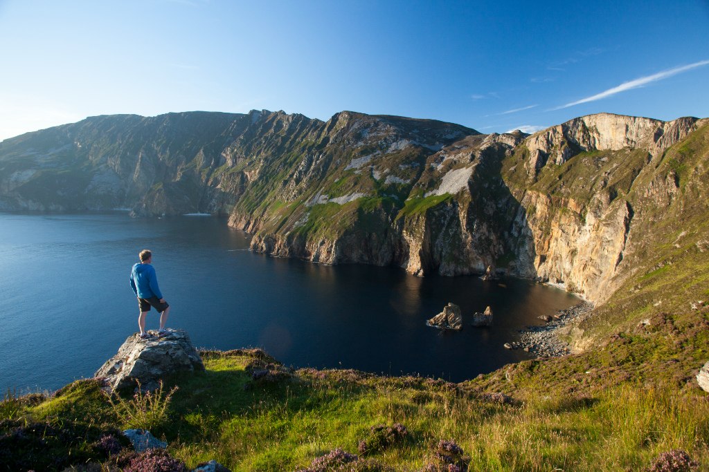 A hiker stands above the towering Slieve League Cliffs, gazing over the deep blue sea and rugged coastline.