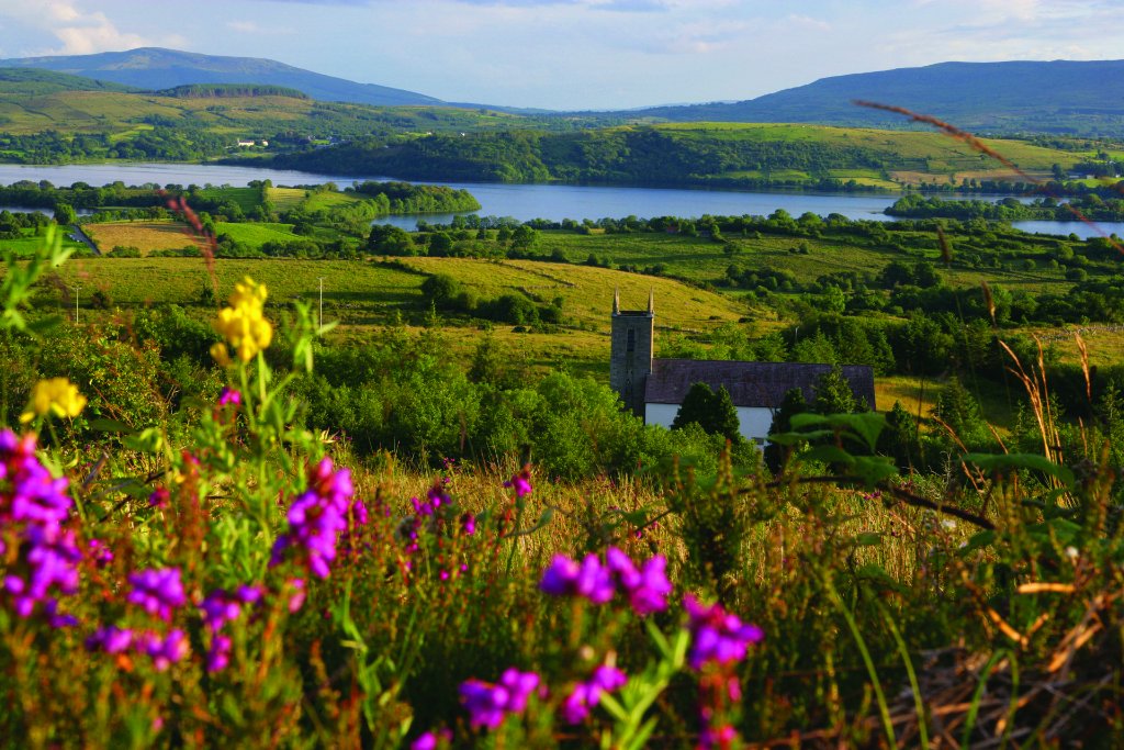 A peaceful lake view with a stone church and rolling hills framed by bright summer wildflowers in the foreground.