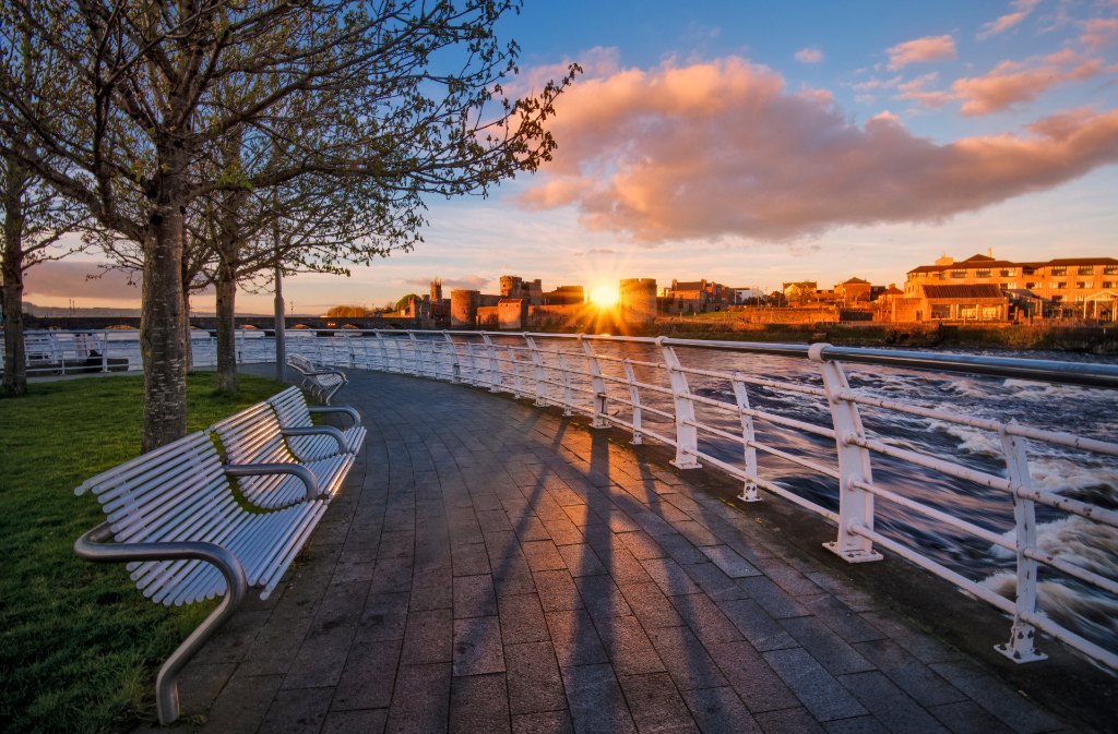 Sunset along Limerick’s riverfront with benches and view of King John’s Castle glowing under golden light.