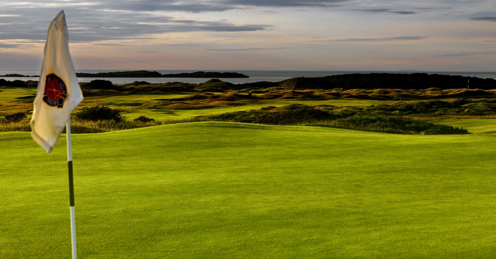 A golf flag stands on lush fairways overlooking the Atlantic Ocean, with golden evening light on rolling dunes.
