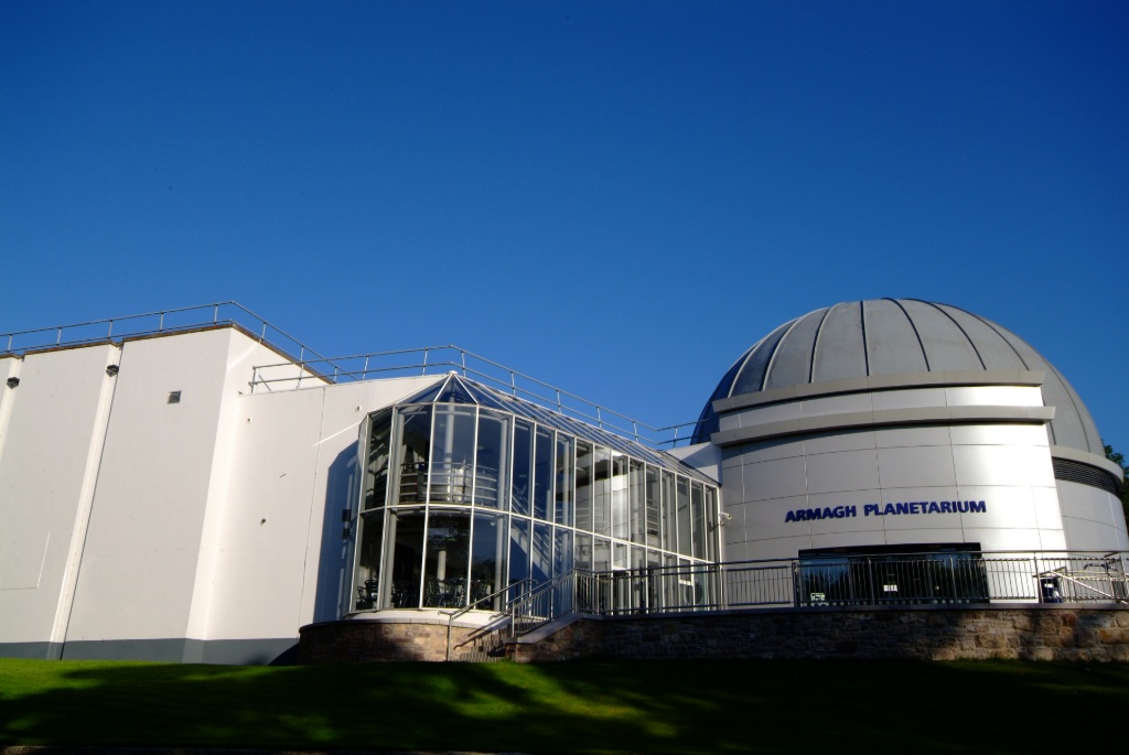 Exterior of Armagh Planetarium with domed roof and glass entrance lit by sunlight under a clear blue sky.