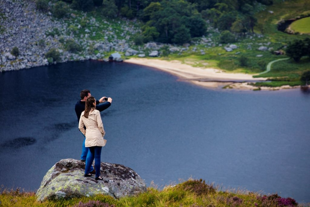 A couple stands on a rock overlooking Lough Tay in County Wicklow, taking in the scenic lakeside view.