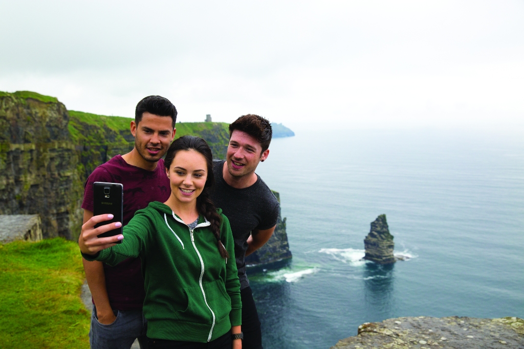 Three friends take a selfie at the edge of the Cliffs of Moher, with sheer sea cliffs rising from the Atlantic below.