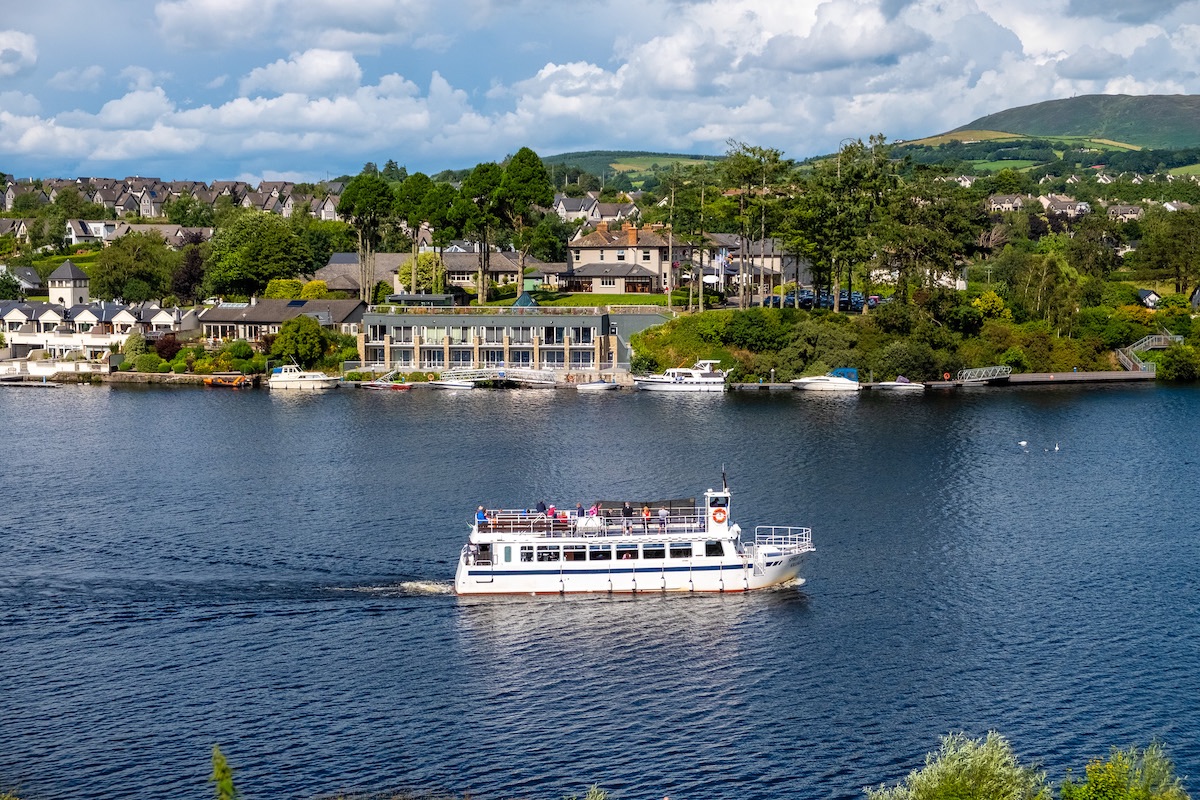 White river cruiser with passengers sailing past leafy riverside homes on Lough Derg, Clare.