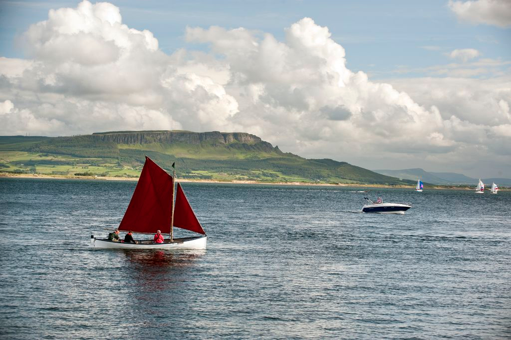 Traditional sailing boat with red sail crossing Donegal Bay beneath mountains on the horizon.