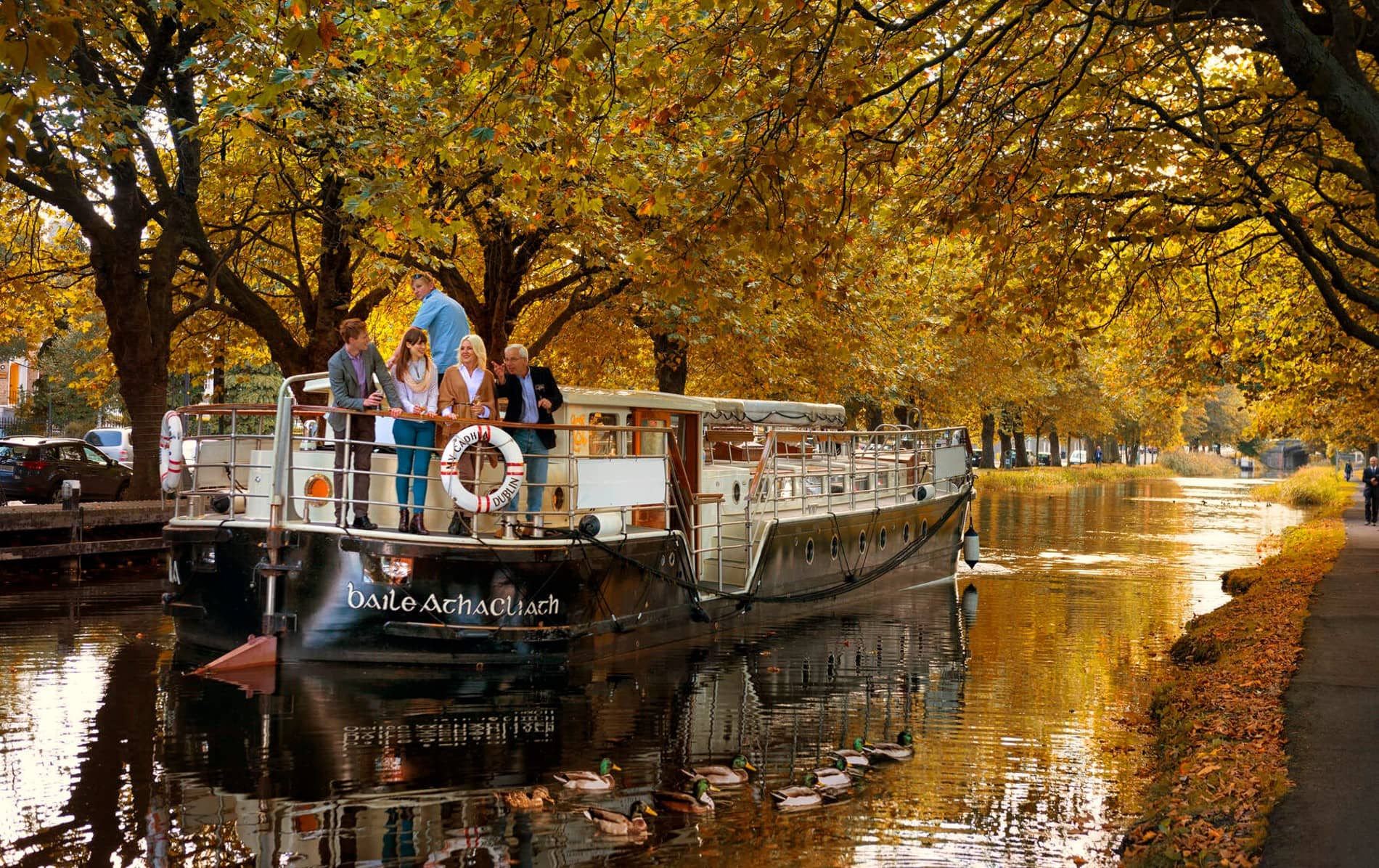 Group of people on a canal boat in Dublin, surrounded by autumn trees and ducks swimming nearby.