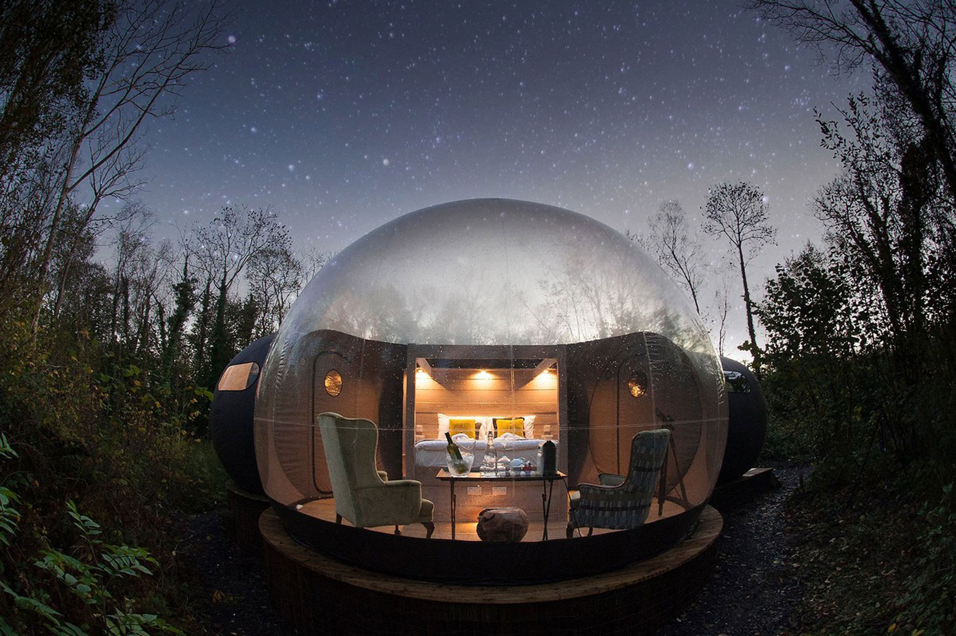 Transparent bubble dome glamping pod set in woodland at Finn Lough, County Fermanagh, with interior warmly lit under a starlit sky.
