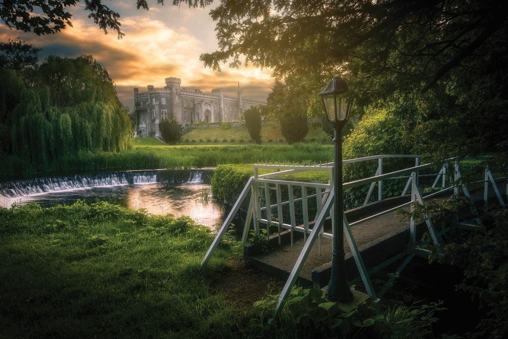 White footbridge over a stream with a view of Bellingham Castle and sunset glow in County Louth.