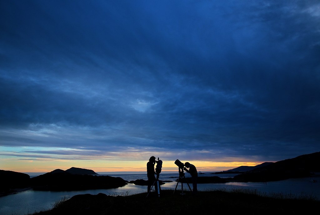 Silhouettes of people stargazing with telescopes against a deep blue twilight sky above rugged coastal mountains.