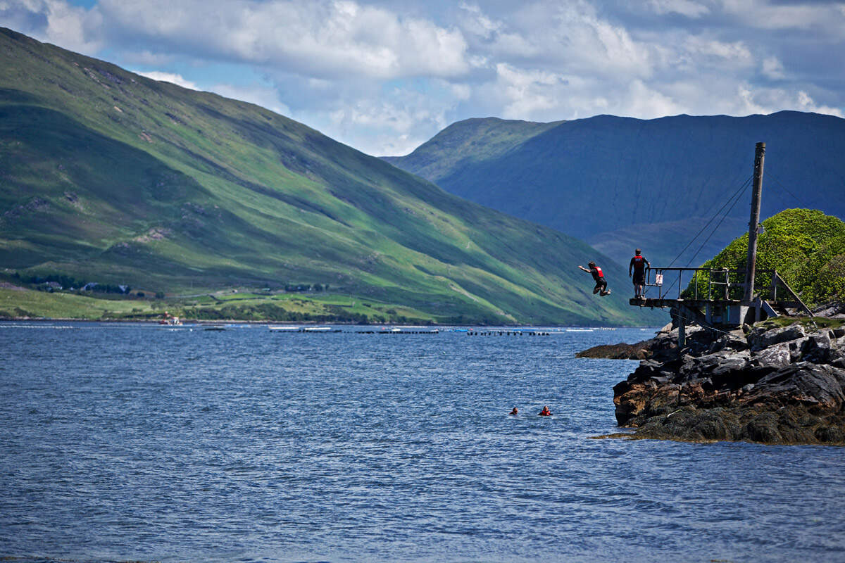 Two people jump off a wooden platform into Killary Fjord, with steep green mountains rising behind in County Galway.