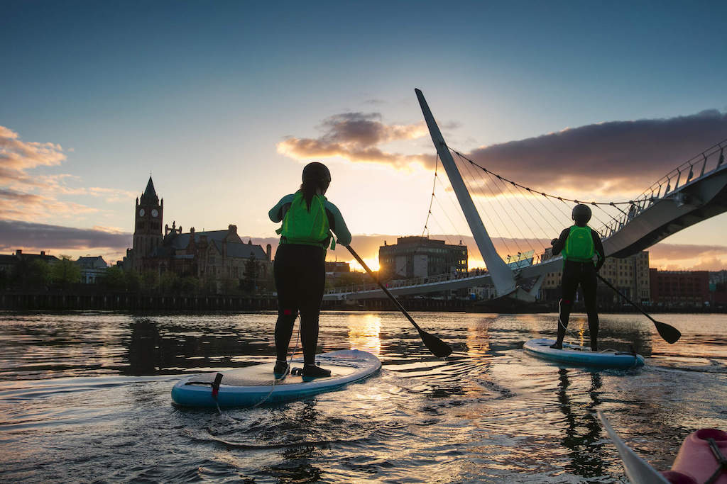 Two paddleboarders at sunset on the River Foyle pass under Derry~Londonderry’s Peace Bridge with Guildhall in view.