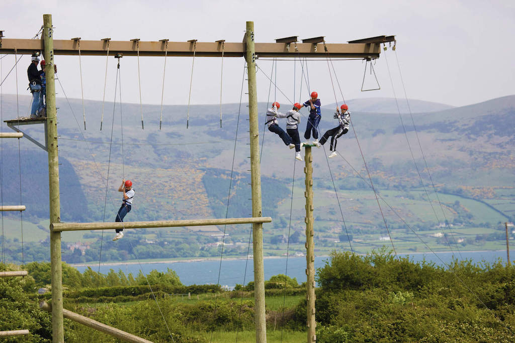 Teens in helmets balance on a high ropes challenge course overlooking Carlingford Lough and the Mourne Mountains.