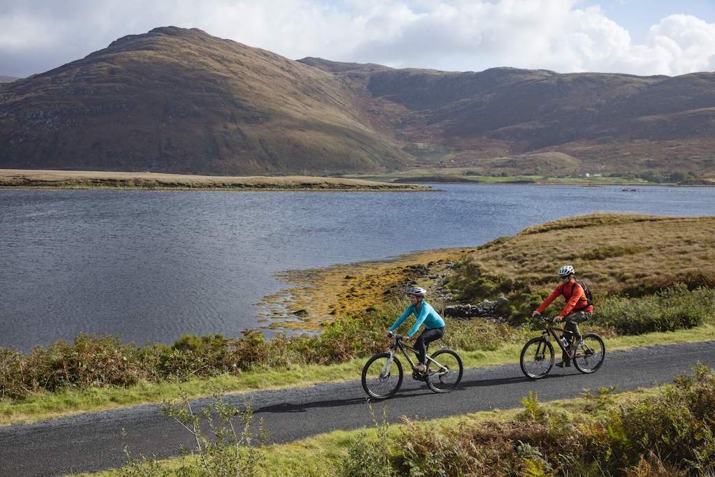 Two cyclists ride beside a tranquil lake surrounded by rugged mountains on the Great Western Greenway, County Mayo.