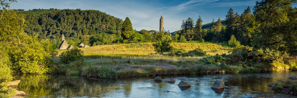 Glendalough Lake Walk in County Wicklow with monastic round tower rising above trees beside a calm river.