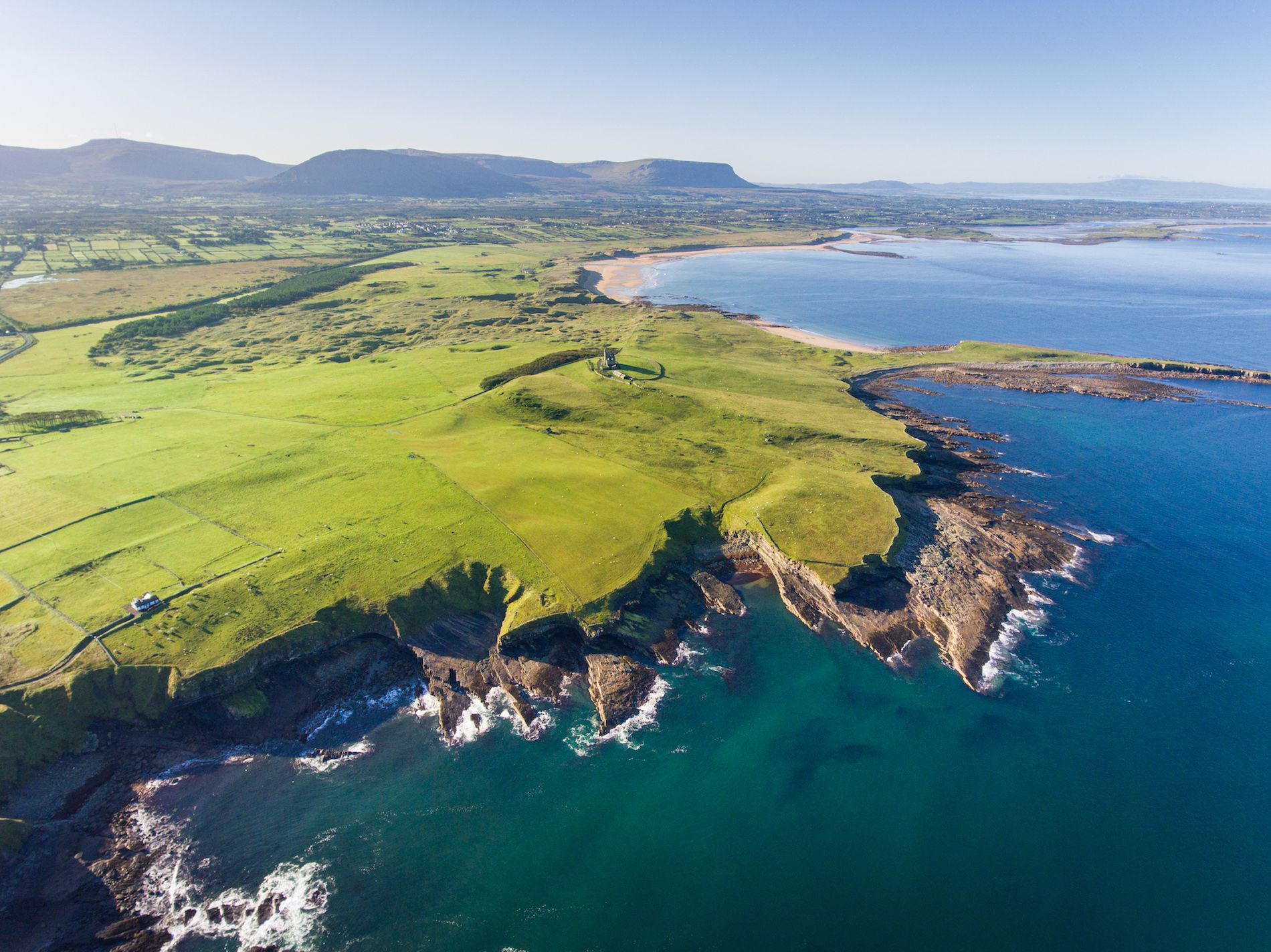 Aerial view of rugged Mullaghmore Head in Sligo, with lighthouse and green fields beside blue Atlantic waters.