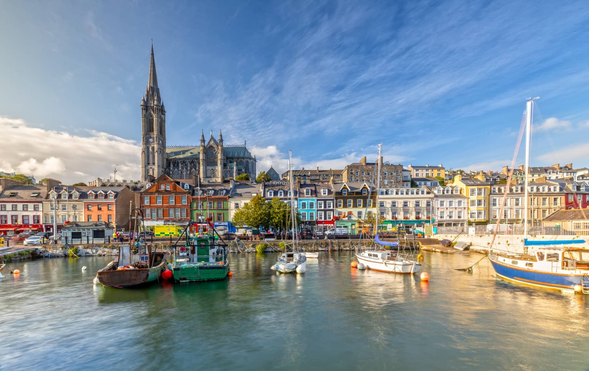 Colourful harbourfront buildings and St Colman’s Cathedral in Cobh, County Cork, viewed from the water on a sunny day.