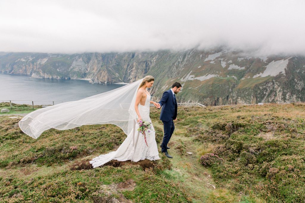 Bride and groom walking along Slieve League Cliffs in Donegal, with ocean and mist-covered cliffs in the background.