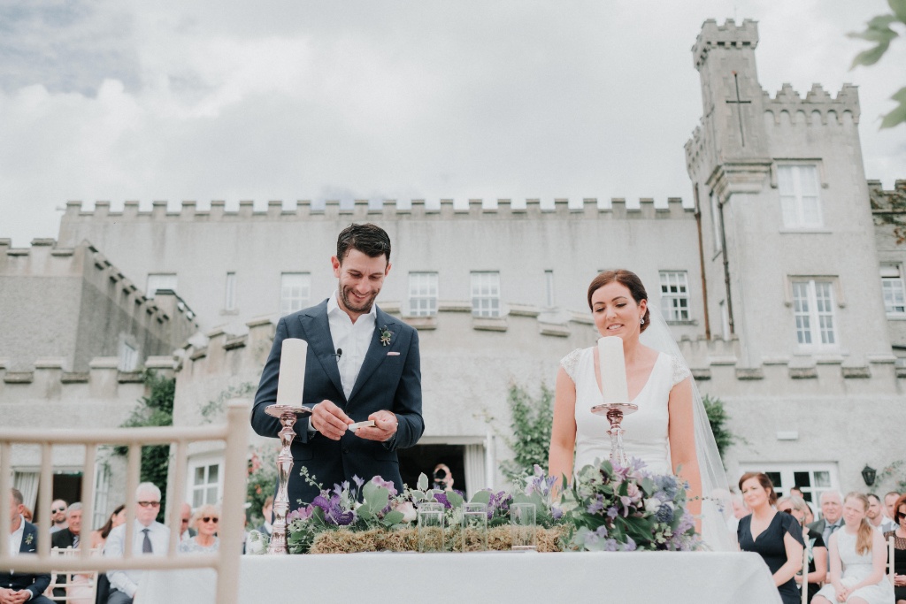 Couple lighting a unity candle during outdoor wedding ceremony at Bellingham Castle in County Louth, with guests seated behind.