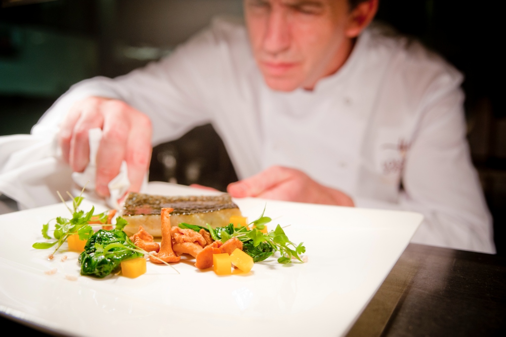 Chef adding final touches to a plated dish at Lough Erne Resort in Fermanagh, garnished with greens and chanterelle mushrooms.