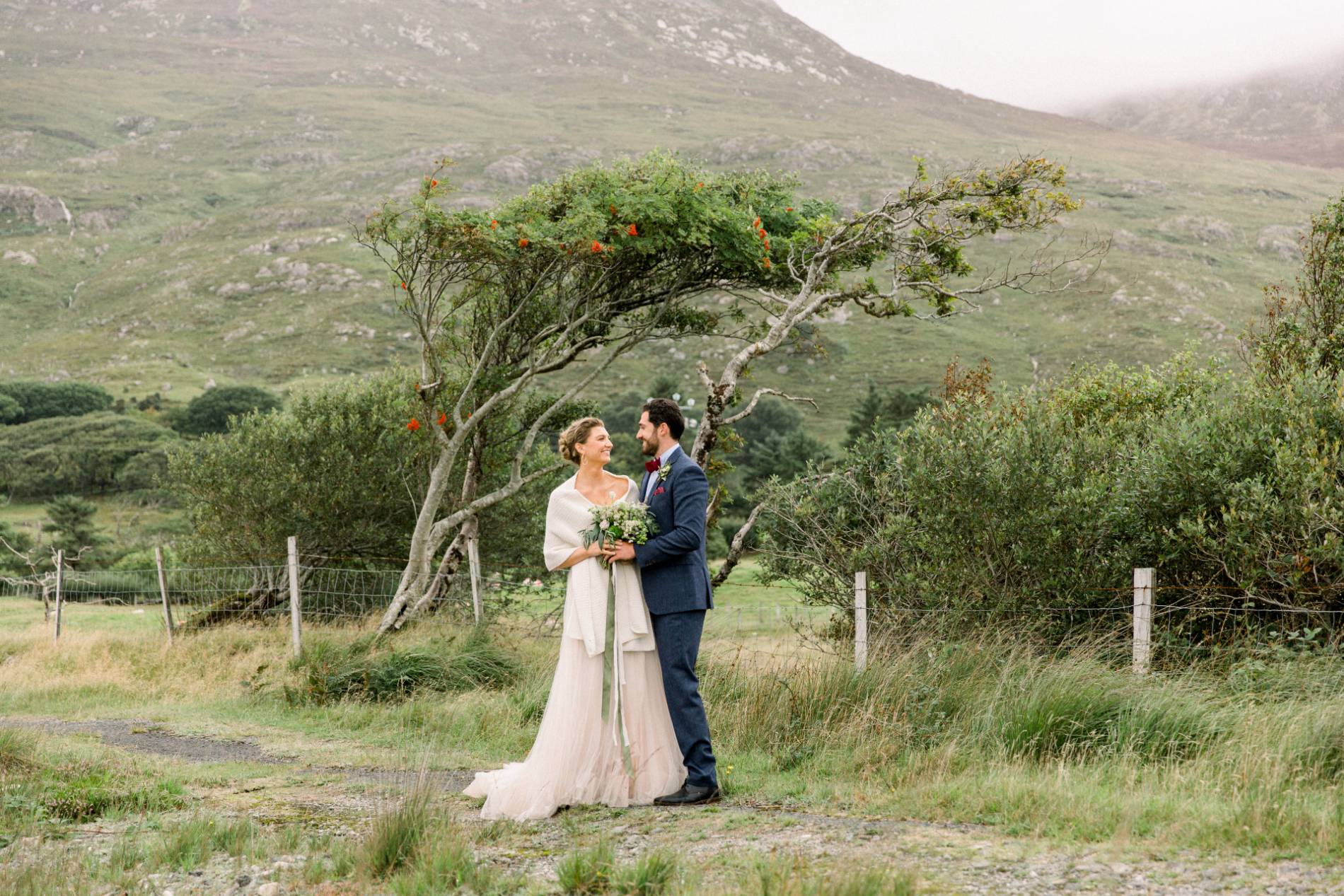 Couple embracing beneath a windswept tree in a rural Irish landscape, symbolising a traditionally Irish wedding celebration.