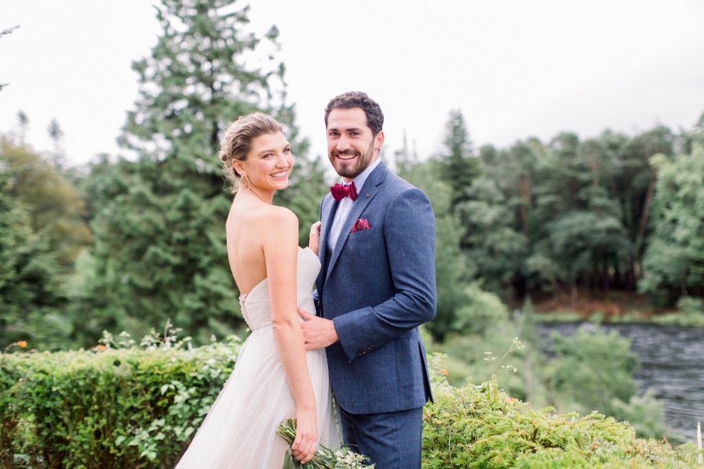 Bride and groom standing arm in arm at Ballynahinch Castle in County Galway, smiling in front of river and forested backdrop.