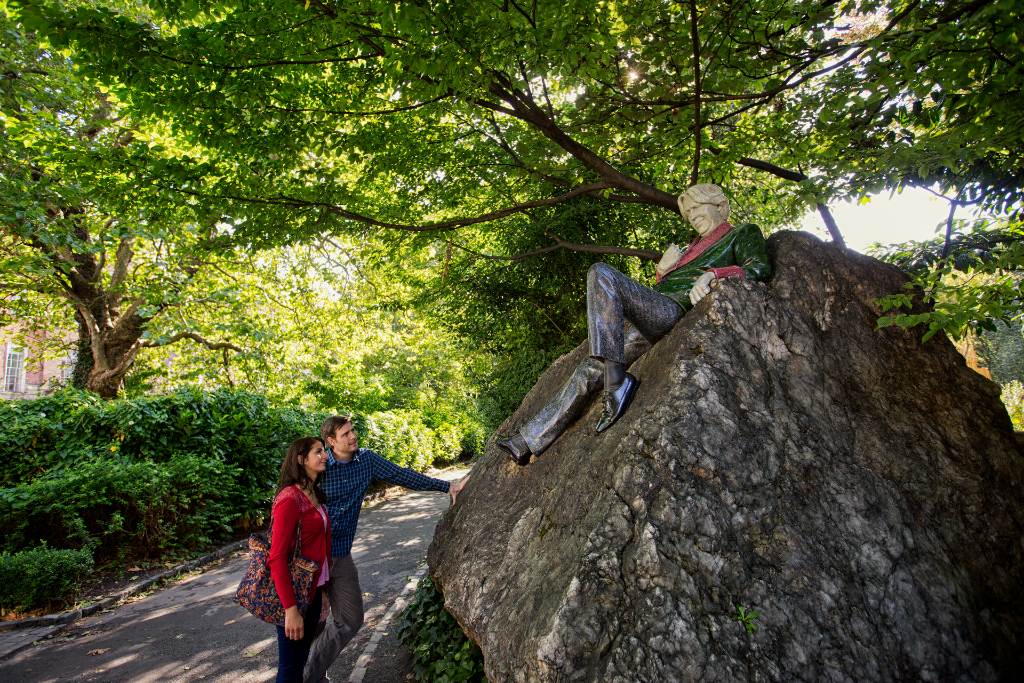 Couple gazes at Oscar Wilde statue in Merrion Square, surrounded by trees and rich greenery.