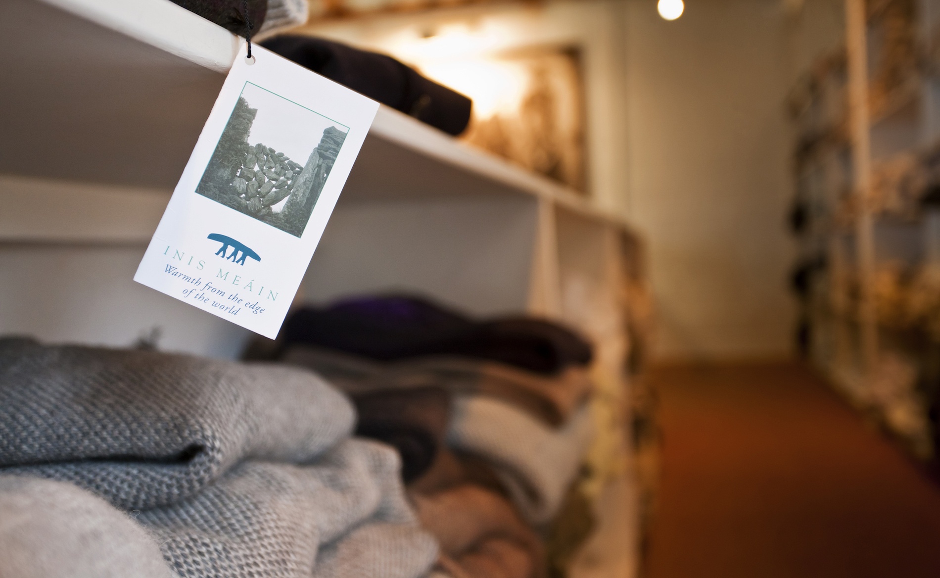 Close-up of an Inis Meáin knitwear tag hanging in a shop with folded Aran jumpers in the background.