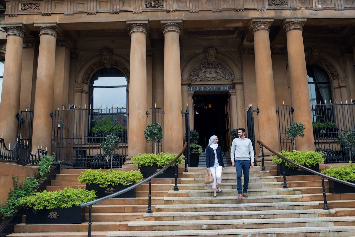 Couple walking down steps of Belfast’s Merchant Hotel, a grand historic building with columns.