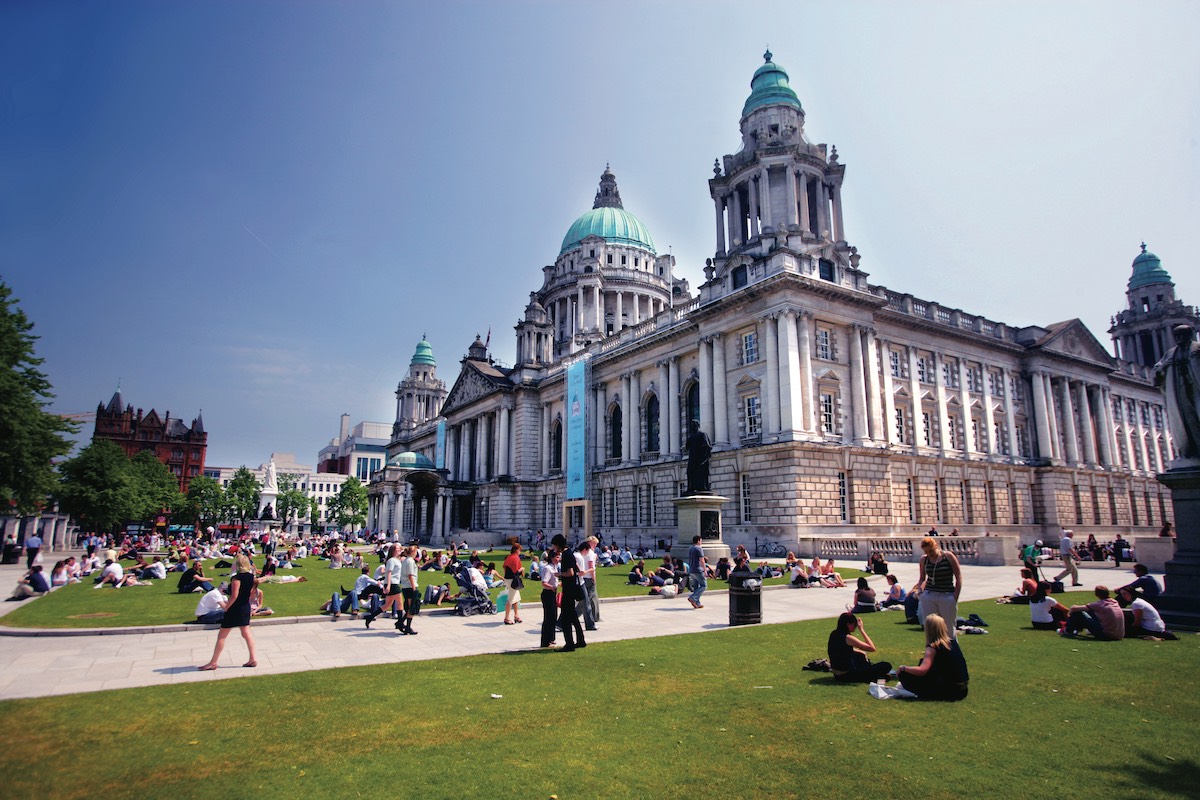 People relaxing on the lawn outside Belfast City Hall on a sunny day with the landmark’s domes and towers in view.