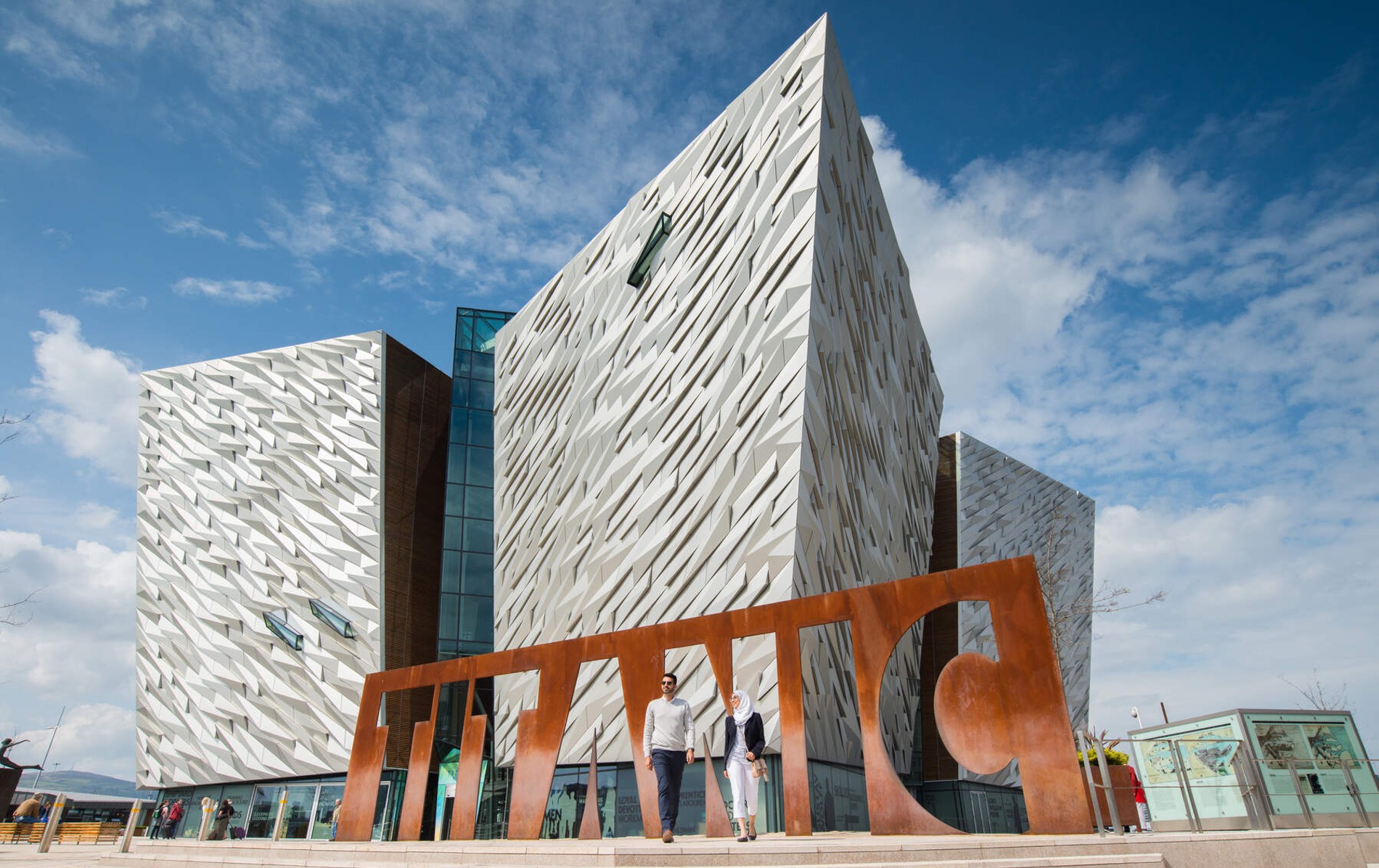 Two visitors walk beside the rusted TITANIC sign outside the bold aluminium-clad Titanic Belfast building.