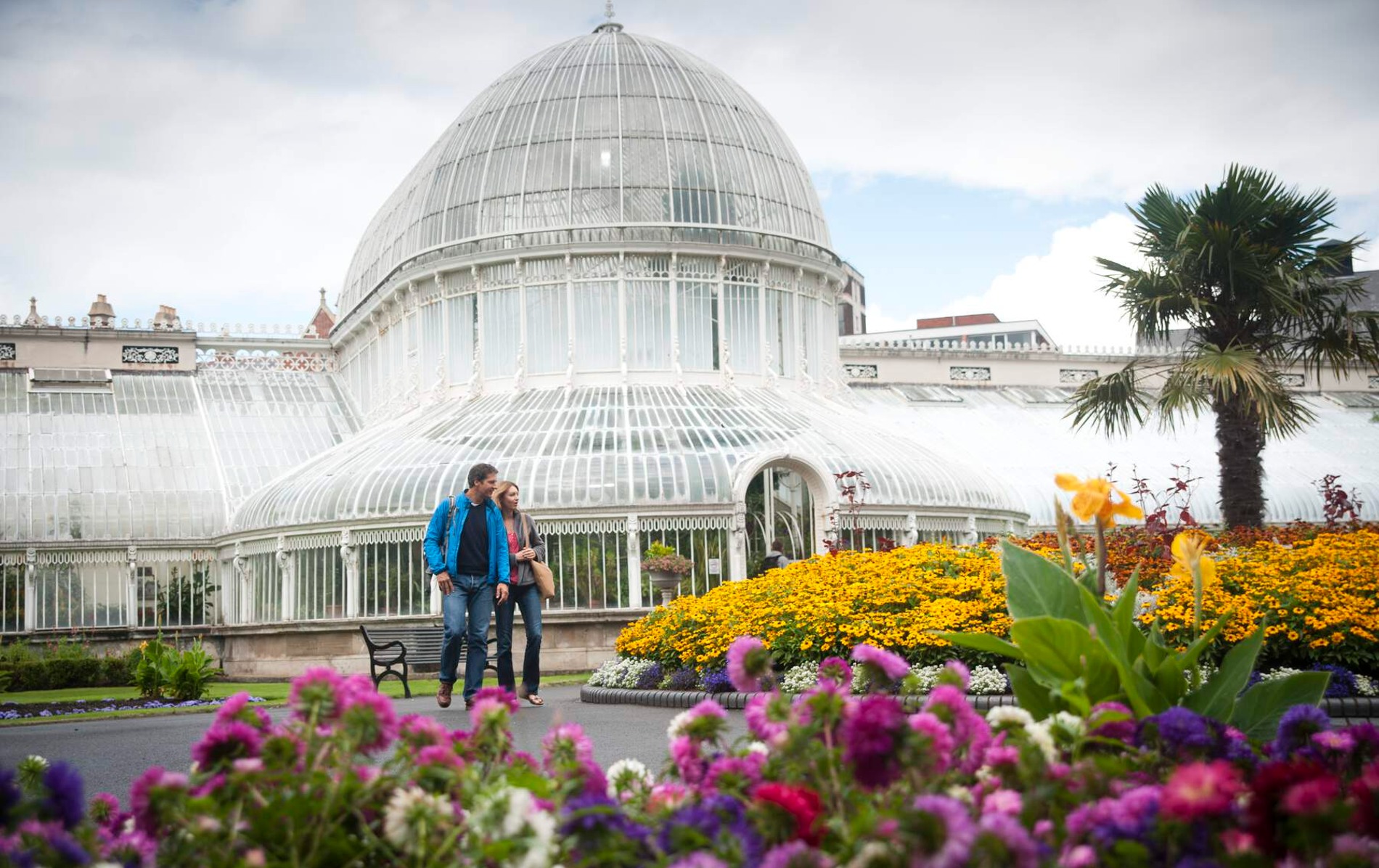Couple walks past flowerbeds and palm trees in front of the domed glass Palm House in Belfast Botanic Gardens.