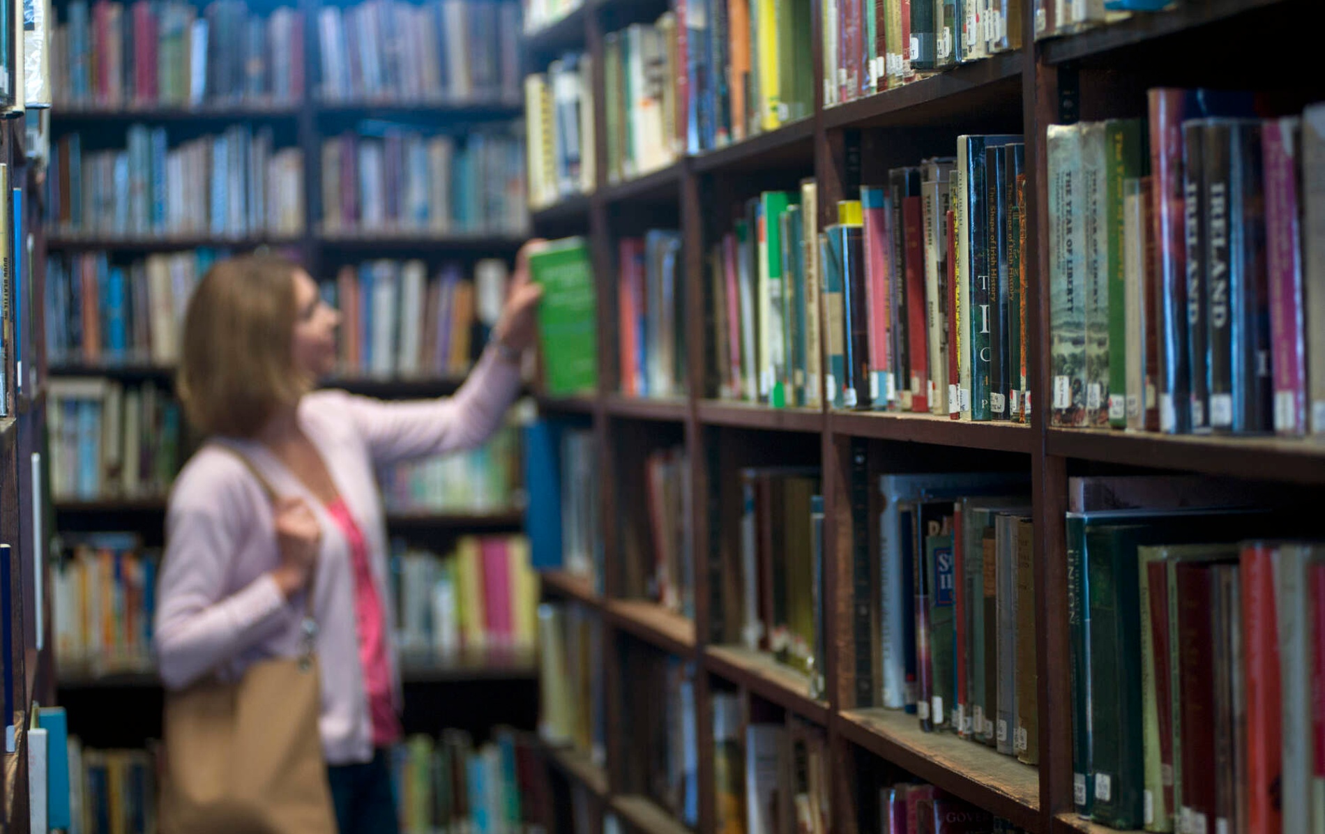 Woman browses a book on a tall shelf in a quiet Belfast library lined with colourful, tightly packed books.