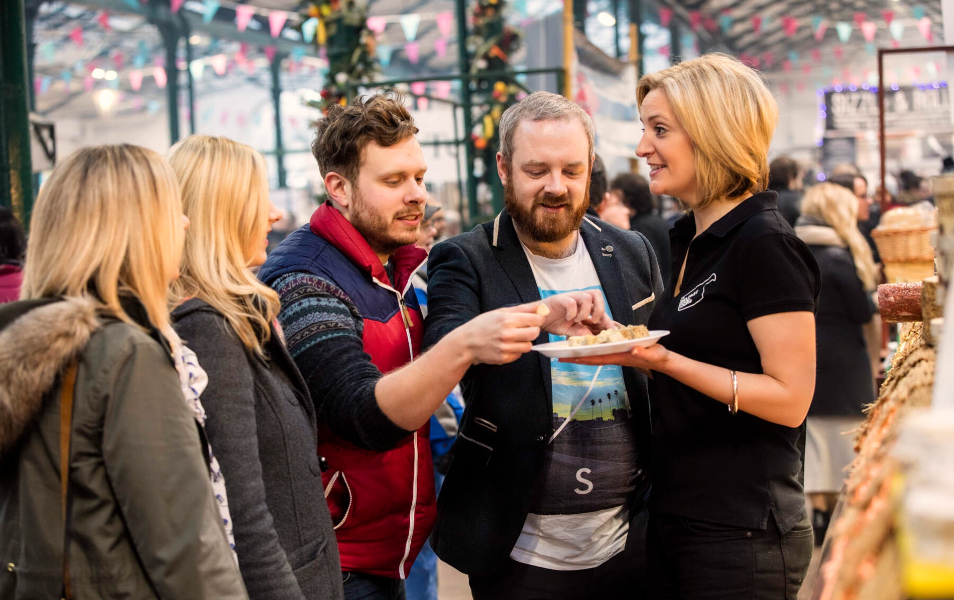People sample food from a smiling vendor at a busy stall in St George’s Market, decorated with bunting.