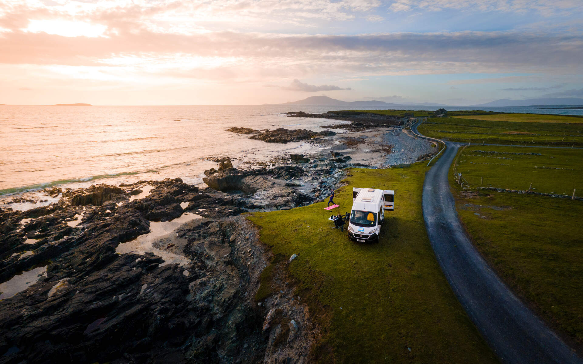 A campervan parked beside a winding coastal road in Ireland, with ocean waves, rocky shore, and a golden sunset sky.