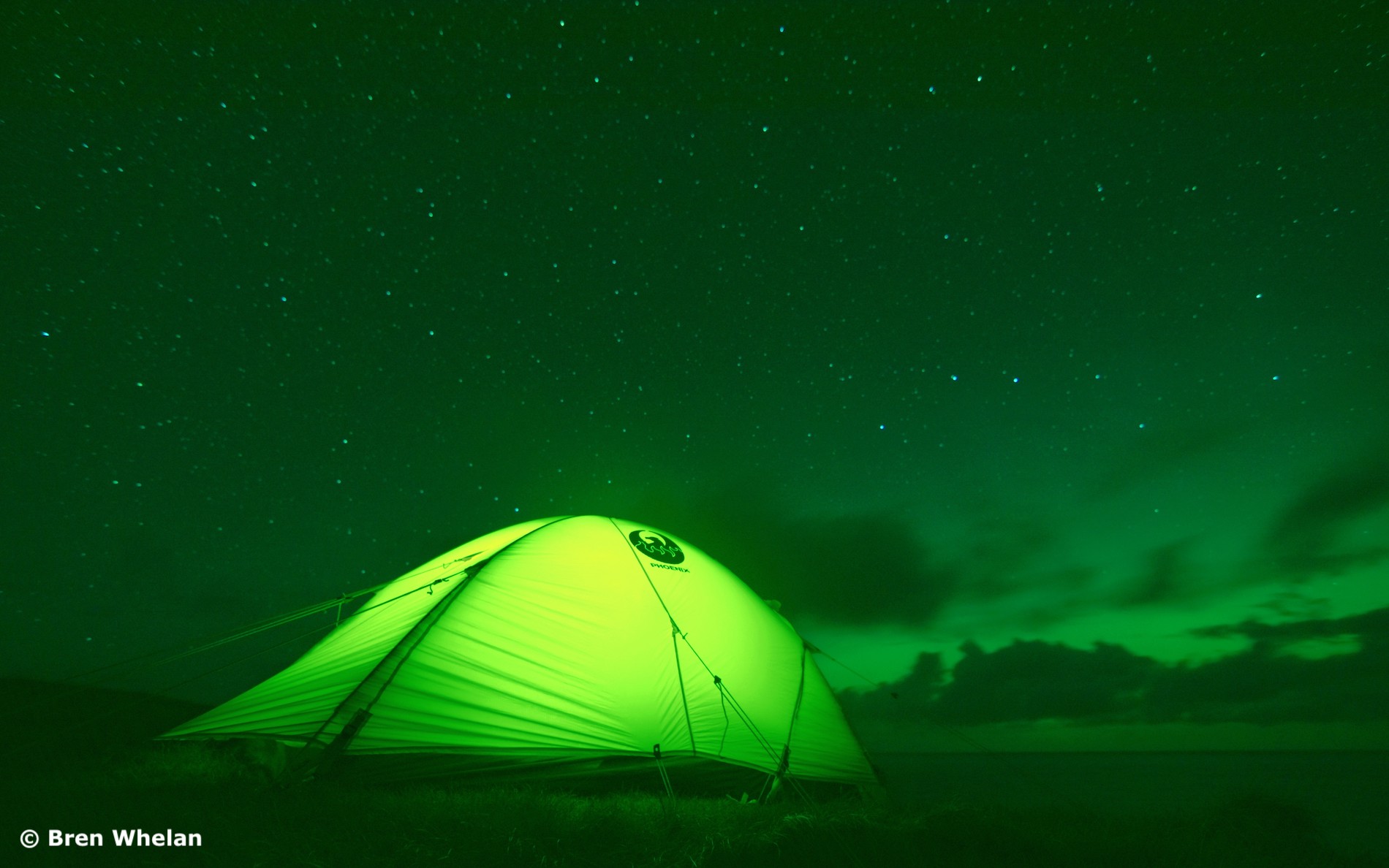 Green tent glows beneath a starry night sky as the Aurora Borealis shimmers above Malin Head’s coastal landscape.