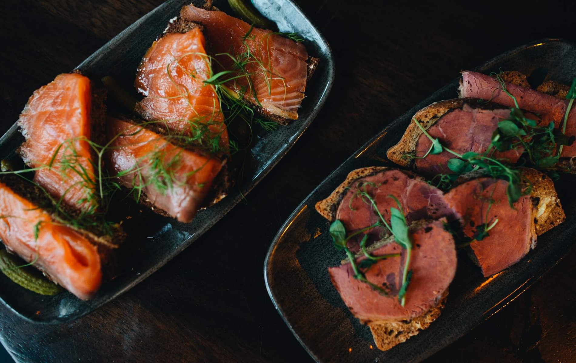 Plates of smoked salmon on rye bread and roast beef on brown bread, garnished with herbs and served as appetisers.