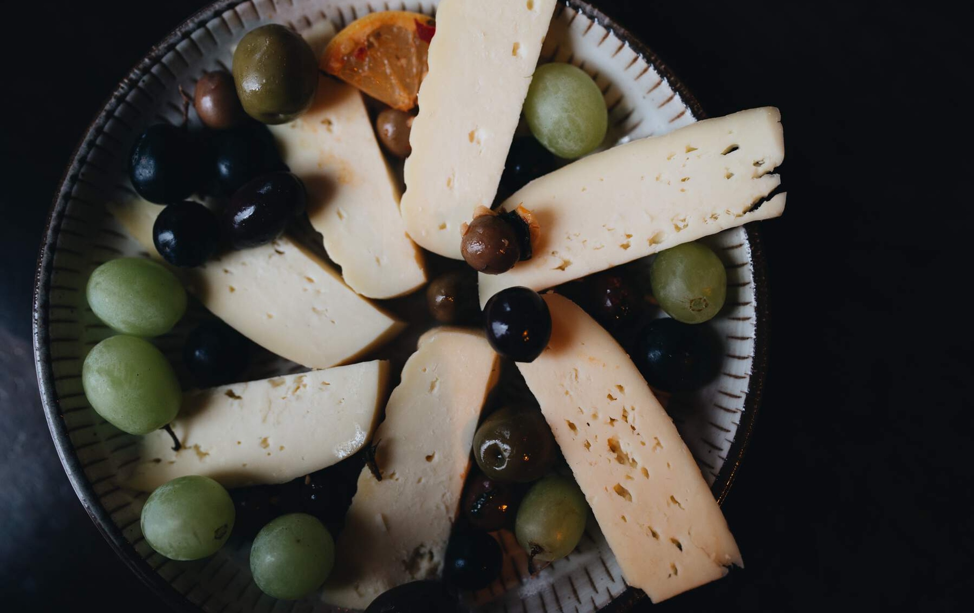Cheese platter with sliced semi-hard cheese, green and black grapes, olives and orange on a decorative plate.