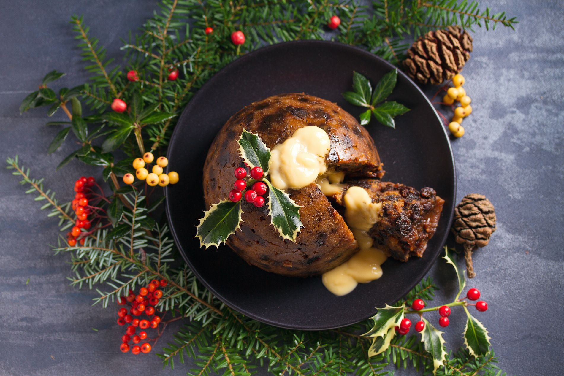 Traditional Irish Christmas pudding with holly garnish and custard, served on a black plate with festive greenery.