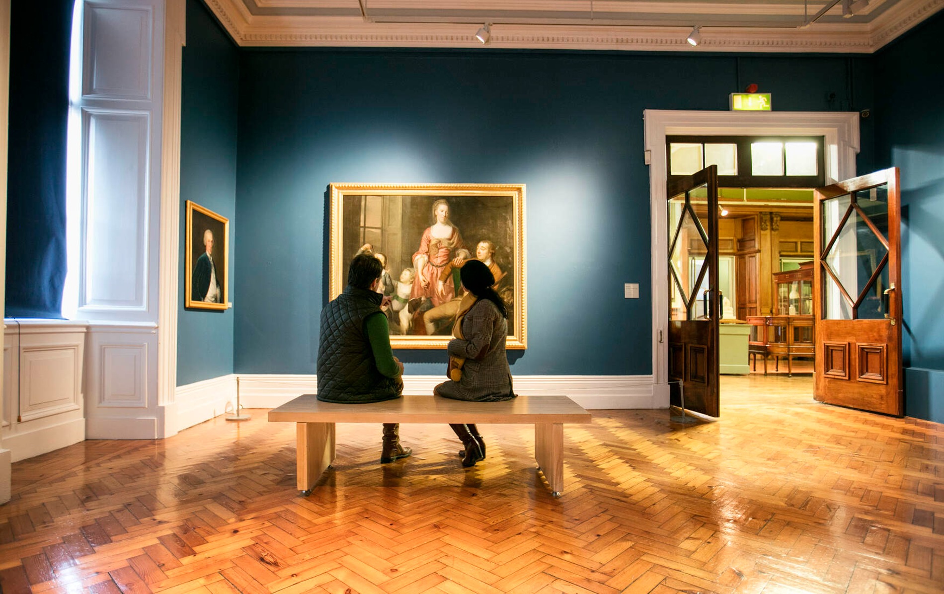 Two visitors seated on a bench admiring a portrait in Crawford Art Gallery, Cork.
