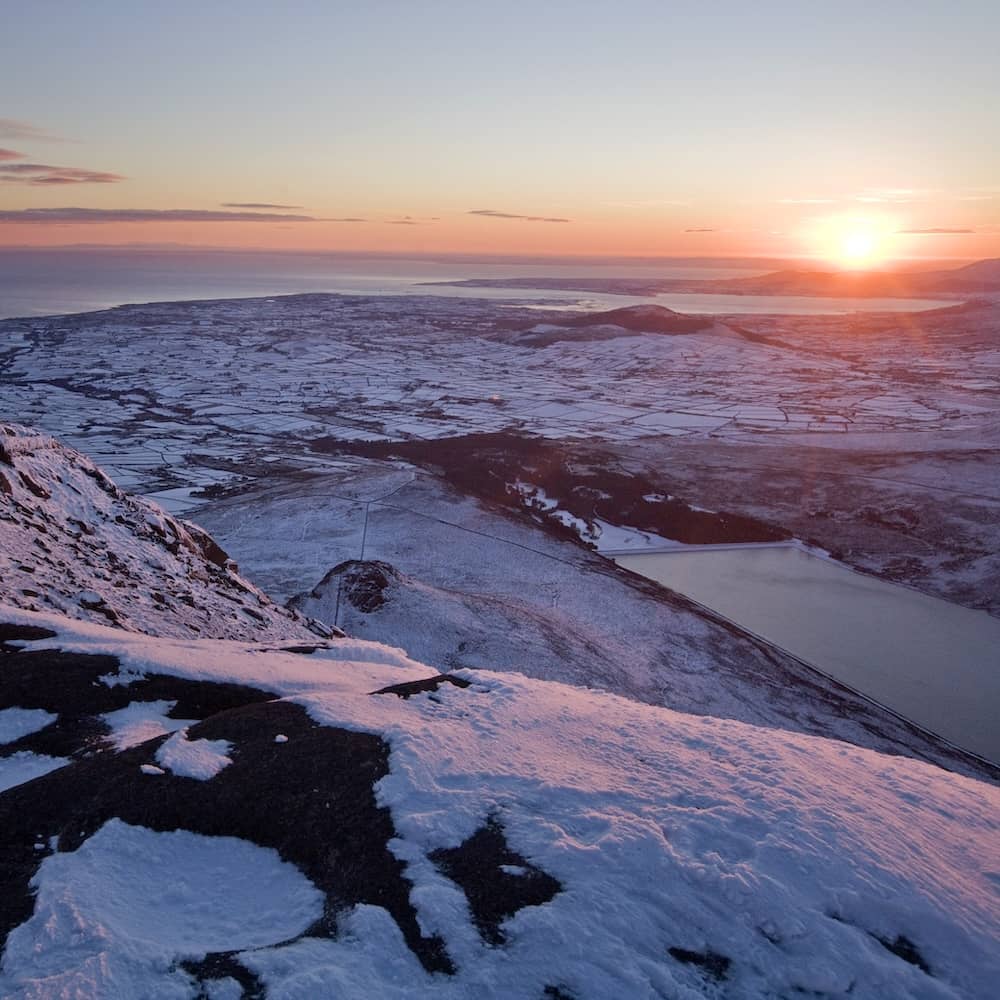 Snow-covered Mourne Mountains at sunrise overlooking rolling countryside, lakes and the coastline near Newry.