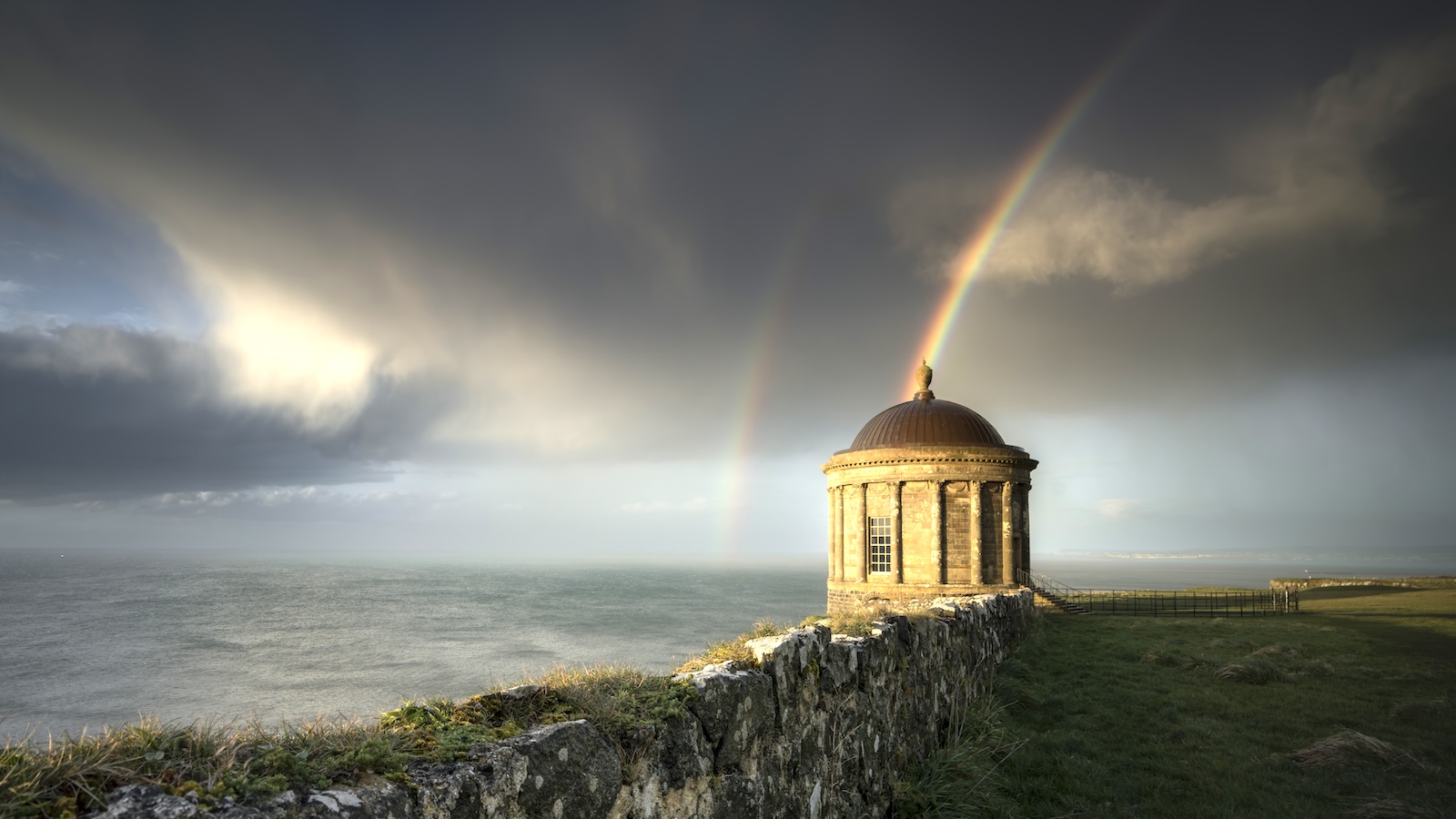 Mussenden Temple in County Londonderry framed by double rainbow above dramatic coastal cliffs.