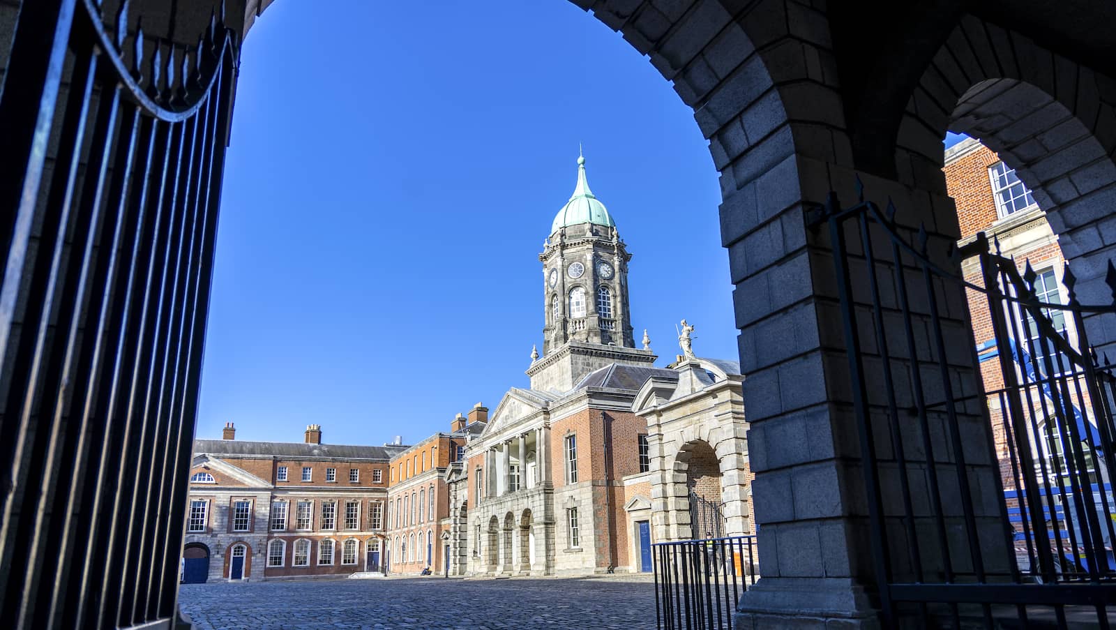 Clock tower and courtyard of historic Dublin Castle viewed through iron gates on a clear day with blue skies.