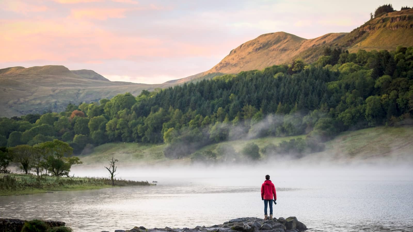 Person in a red coat standing by the misty shores of Glencar Lake in Leitrim, surrounded by forested hills at sunrise.