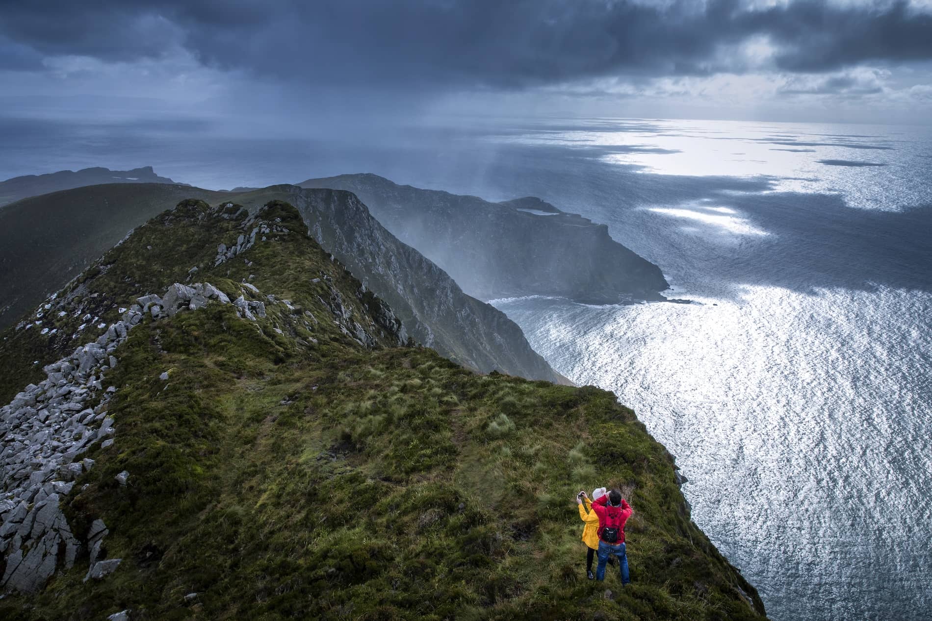 A couple takes a photo on Slieve League, with dramatic cliffs and the Atlantic Ocean under a brooding sky.