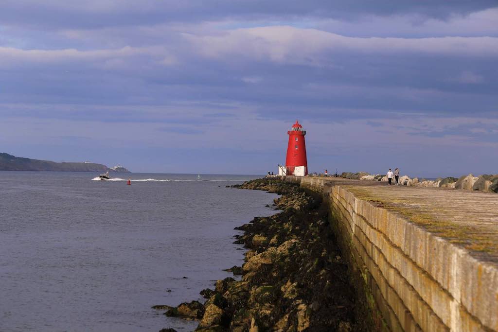 People walk the long stone pier toward the red Poolbeg Lighthouse, with a powerboat speeding through the bay nearby.