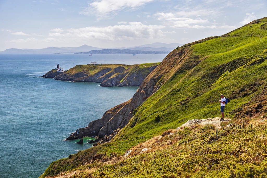 A hiker stands on a grassy cliff path overlooking the sea and lighthouse at Howth Head on a sunny day.