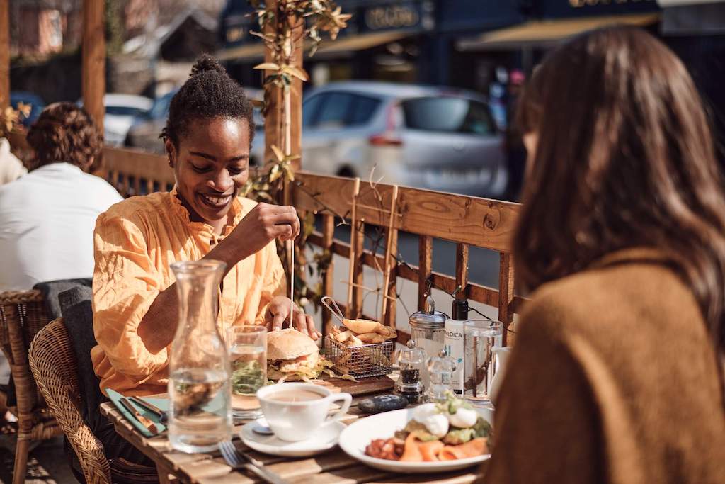 Two women enjoy brunch and coffee at a sunny outdoor café table in Dalkey, with plates of fresh food and relaxed smiles.